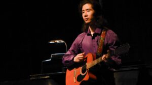 A student playing guitar and singing at a recital